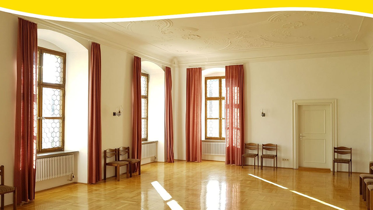 Großer, heller Barocksaal mit glänzendem Parkettboden und roten Vorhängen. Am oberen Bildrand verläuft eine gelbe Design-Welle.